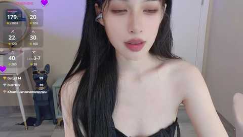 Media: Video of a young, slim Asian woman with long black hair, wearing a black strapless top, eyes closed, in a modern room with a blue bag and a white door in the background.