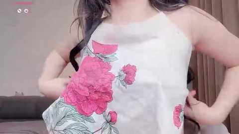 Media: A video of a light-skinned woman with long dark hair, wearing a sleeveless white top with large pink floral patterns. She stands indoors with a brown curtain in the background.