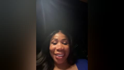 Media: Video of a smiling Black woman with long, dark hair, wearing a blue top, captured in a dimly lit room.