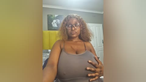 Media: Video of a dark-skinned woman with curly hair, wearing glasses and a gray tank top, holding her breasts. She's in a bedroom with a yellow headboard and a framed landscape painting on the wall.