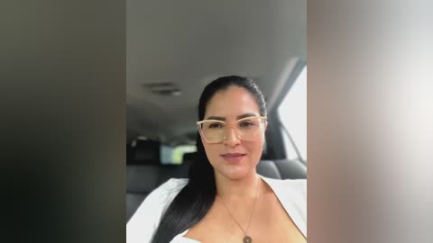 Media: Video of a light-skinned woman with long black hair, wearing glasses and a white lab coat, seated in a car, with blurred background.