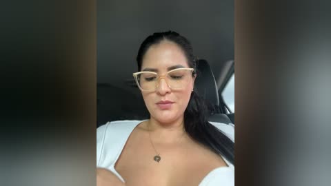 Media: Video of a Latina woman with olive skin, long dark hair, wearing glasses, a white blouse, and a necklace, seated in a car, looking down with a neutral expression.