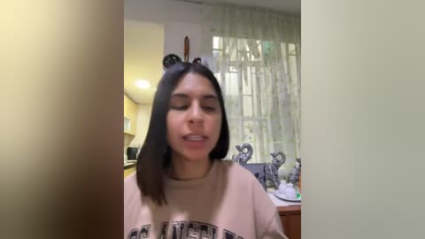 Media: A candid photo of a young woman with straight black hair, wearing a light beige t-shirt, standing indoors in a kitchen with light green walls and a window.