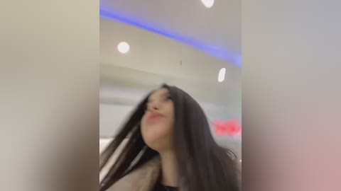 Media: Video of a young woman with long black hair, fair skin, and full lips, looking upward, in a modern, brightly lit room with recessed lights and blue LED strip lighting.