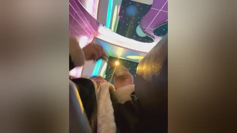 Media: Video of a person in a dark room, illuminated by a glowing yellow light, wearing a white sweater, with a neon-lit futuristic ceiling featuring geometric patterns and stars.