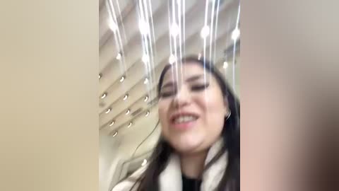 Media: Video of a smiling woman with long dark hair, wearing a white and black scarf, captured from a low angle with a reflective ceiling showing numerous lights.