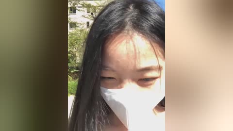 Media: A close-up video of an Asian woman with long black hair, wearing a white surgical mask, looking downward. The background features a blurred, outdoor setting with green foliage and a building. The image has a soft focus.