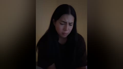 Media: A dimly lit video of a young woman with long, straight black hair and fair skin, wearing a black shirt, standing in a dark, narrow hallway with beige walls. Her expression is serious and focused.