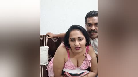 Media: A video showing a middle-aged South Asian couple in a dimly lit room; woman with black hair, pink dress, and medium breast size; man with dark hair, mustache, and white shirt.