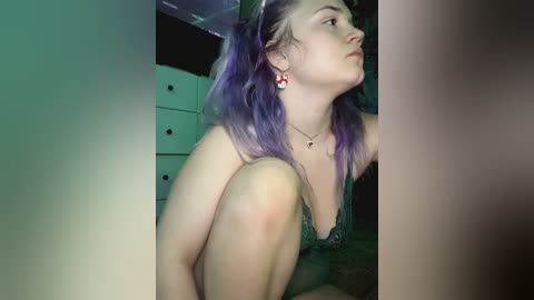 Media: Video of a young woman with light skin and purple hair, wearing a green lace bra and red earrings, sitting on a bed in a dimly lit room with a white dresser in the background.