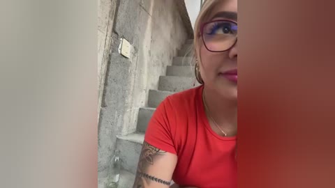 Media: Video of a smiling, fair-skinned woman with glasses, red t-shirt, and arm tattoos, standing in a narrow, concrete stairwell.