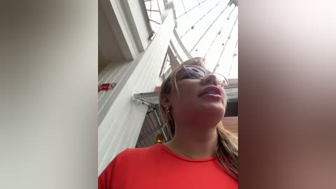 Media: Video of a smiling, light-skinned woman with straight brown hair, wearing a bright red top, standing in an indoor space with white walls and a large, glass-paned ceiling.