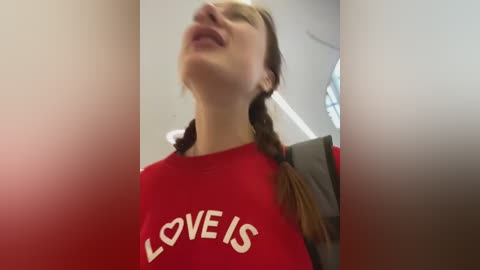 Media: Video of a Caucasian woman with brown hair in braids, wearing a red \"LOVE IS\" t-shirt, leaning back with a content expression, indoors with blurred background.