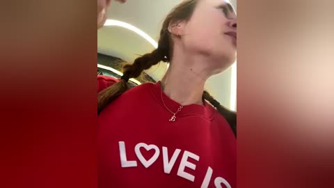 Media: Video of a young woman with light skin, brown hair in braids, wearing a red \"LOVE\" sweatshirt, standing indoors with a blurred background.