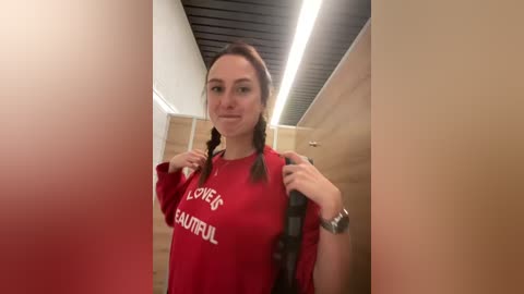 Media: Video of a young woman with light skin, brown hair in braids, wearing a red \"LOVER FAULTY\" T-shirt, standing in a beige-tiled bathroom with a black ceiling.