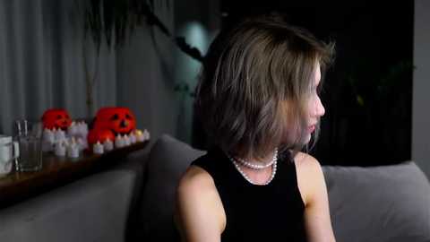 Media: Video of a woman with short, dark hair, wearing a black sleeveless top and pearl necklace, seated on a gray couch. Background includes a shelf with three red pumpkin-shaped decorations and a green plant.