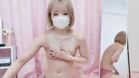 Media: A video of a topless Asian woman with fair skin and a short blonde bob, wearing a white mask, standing in front of a mirror.