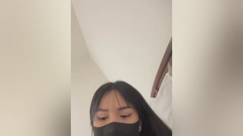 Media: A video of an Asian woman with long black hair, wearing a black face mask, standing in a narrow hallway with white walls and a wooden door.