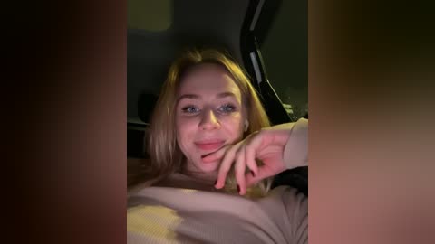 Media: A video of a young Caucasian woman with fair skin and long blonde hair, smiling slightly, wearing a beige sweater. The background is dimly lit, suggesting a car interior, with a steering wheel partially visible.