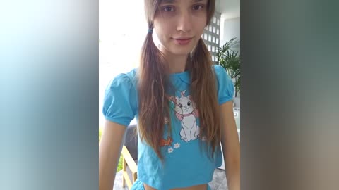 Media: Video of a young, fair-skinned woman with long brown pigtails, wearing a blue crop top featuring a cartoon rabbit design, indoors with blurred background.