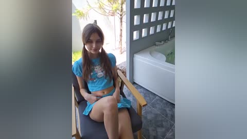 Media: Video of a young woman with long brown hair, wearing a blue crop top and matching shorts, seated on a wooden chair in a modern bathroom.