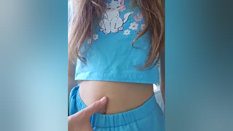 Media: Video of a woman with light skin, wearing a blue crop top with a cartoon cat and flowers, and matching blue pants, being touched on her stomach by a hand.