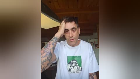 Media: Video of a tattooed man with short dark hair, wearing a white t-shirt with a graphic design, standing indoors with wooden ceiling and dim lighting.