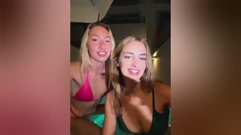 Media: Video of two young women, one with blonde hair in a pink bikini, the other with brown hair in a green top, smiling, indoors with a dimly lit background.