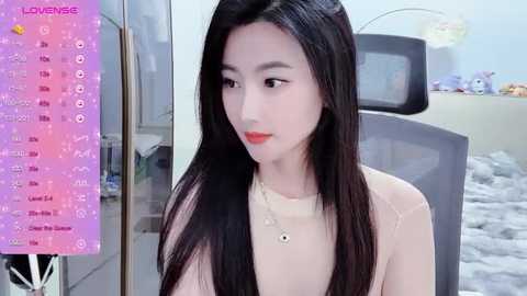 Media: A video of a young East Asian woman with long black hair, fair skin, and red lipstick, wearing a beige sweater, sitting in a modern room with a gray chair.