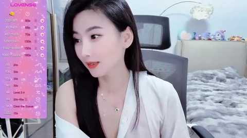 Media: Video of an East Asian woman with long black hair, fair skin, and a slender physique, wearing a white blouse. She sits in a modern, light-colored room with a gray chair, a bed, and decorative items.