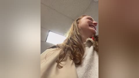 Media: Video of a smiling, Caucasian woman with long, wavy brown hair, wearing a beige coat and a white sweater. She stands in a bright, indoor setting with a ceiling light.