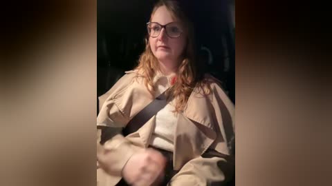 Media: Video of a young woman with long, wavy light brown hair, wearing glasses, a beige trench coat, and a white shirt, sitting indoors, with a dimly lit background.