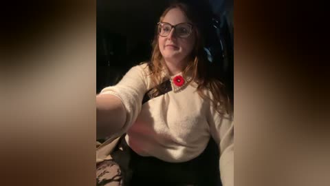 Media: Video of a smiling, fair-skinned woman with glasses, wearing a beige sweater, red pin, and black skirt, driving a car.