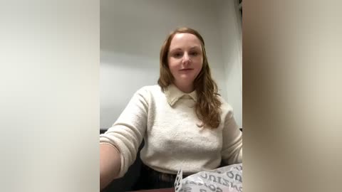 Media: Video of a fair-skinned, red-haired woman in a beige sweater, sitting in an office with a blurred background. She smiles gently, holding a document with text.