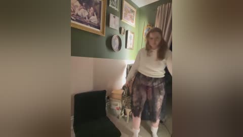 Media: A video shows a young woman with long hair in a white sweater and patterned pants, leaning over a black chair in a dimly lit room with green walls.