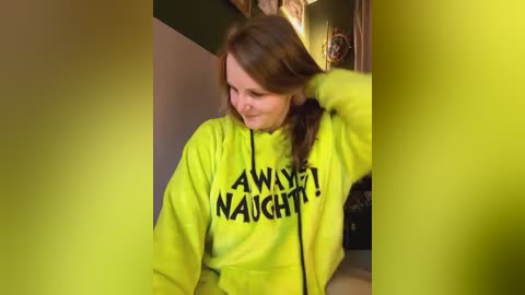 Media: Video of a young woman with light skin and brown hair, wearing a neon green hoodie with the phrase \"AWESTRUCK NAUGHTY\" printed on it. She's indoors, with a warm, yellowish tint in the background.