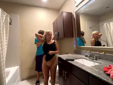 Media: Video of a young blonde woman in a black tank top and beige underwear, adjusting her hair in a bathroom with a dark countertop, white sink, and mirror.