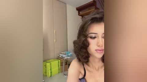 Media: Video of a young woman with curly brown hair, wearing a black bra, standing in a beige room with a green box, wooden shelf, and a closed door.