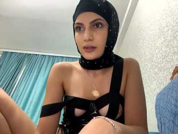 Media: Video of a woman with light brown skin, wearing a black, see-through latex top with straps, and a black hijab adorned with silver studs, in a room with teal curtains and white wallpaper.