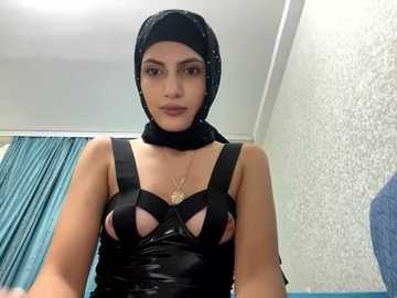 Media: Video of a woman with medium skin tone, wearing a black, strappy, latex top and a black hijab, standing in a room with white textured wallpaper and teal curtains.