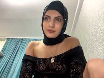 Media: Video of a Middle Eastern woman with light skin, wearing a black hijab, off-shoulder black lace top, and a gold pendant necklace. She's indoors with white walls, teal curtains, and a subtle floral wallpaper.