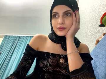 Media: Video of a Middle Eastern woman with medium skin tone, dark eyes, and long black hair under a black hijab, wearing a black lace dress and a necklace, in a room with teal curtains and a floral wallpaper.