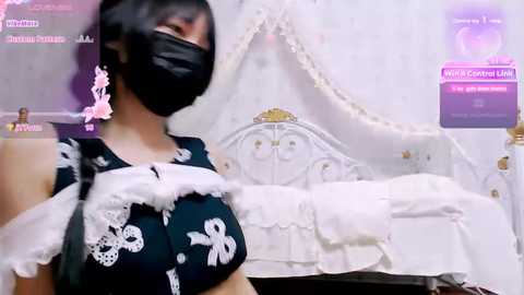 Media: A video of a woman with a light skin tone, wearing a black face mask, black lace dress, and pink earrings, standing in a white, ornately decorated bedroom.