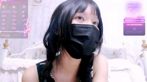 Media: A video of a young Asian woman with long black hair, wearing a black surgical mask, sitting on a white bed with white lace curtains in the background.