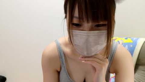 Media: Video of an Asian woman with light skin, brown hair, and bangs, wearing a gray tank top and a white surgical mask, sitting indoors with a colorful, patterned pillow visible in the background.