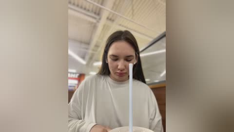 Media: Video of a young woman with long brown hair, wearing a white sweater, eating noodles with a white spoon in a cafeteria with a high ceiling and visible ventilation ducts.