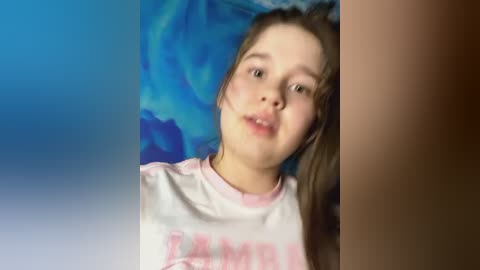 Media: Video of a young Caucasian girl with fair skin and straight brown hair, wearing a white t-shirt with pink text, against a blue and green patterned background.