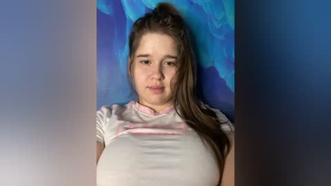 Media: Video of a young Caucasian woman with fair skin, long brown hair, and a neutral expression. She wears a white t-shirt with a pink pattern. The background features a gradient of blue and purple hues.