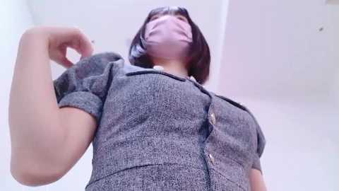 Media: Video of an Asian woman with a medium build, wearing a gray, button-up blouse, pink face mask, and dark hair. She looks up, her arm raised. Background is a white, minimalist room.