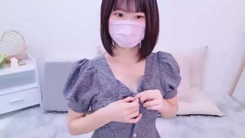 Media: A video of an East Asian woman with a light complexion and shoulder-length black hair, wearing a grey, puffy-sleeved dress and a pink surgical mask, standing on a white fluffy rug in a minimalist room.